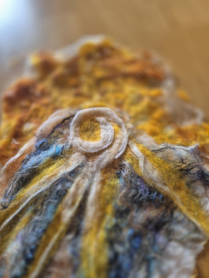 Close-up of a textured, multicolored felted artwork featuring swirling patterns and various colors including yellow, blue, black, and shades of brown.