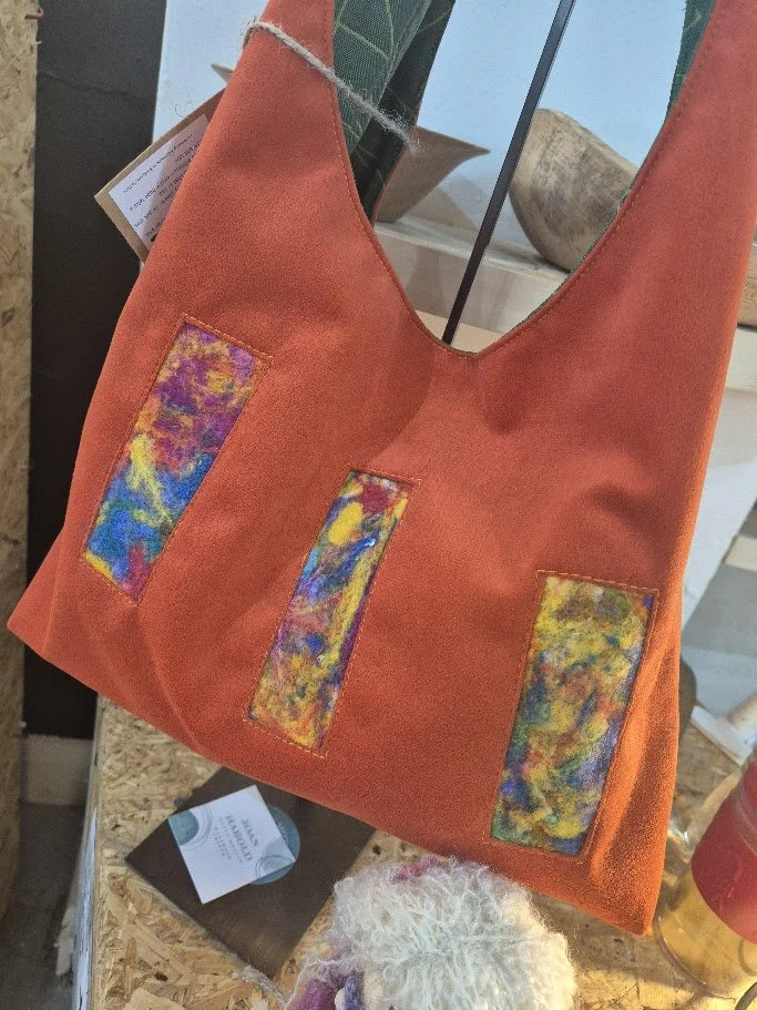 An orange tote bag with three vertical fabric patches that have a colorful abstract pattern, displayed inside a store.