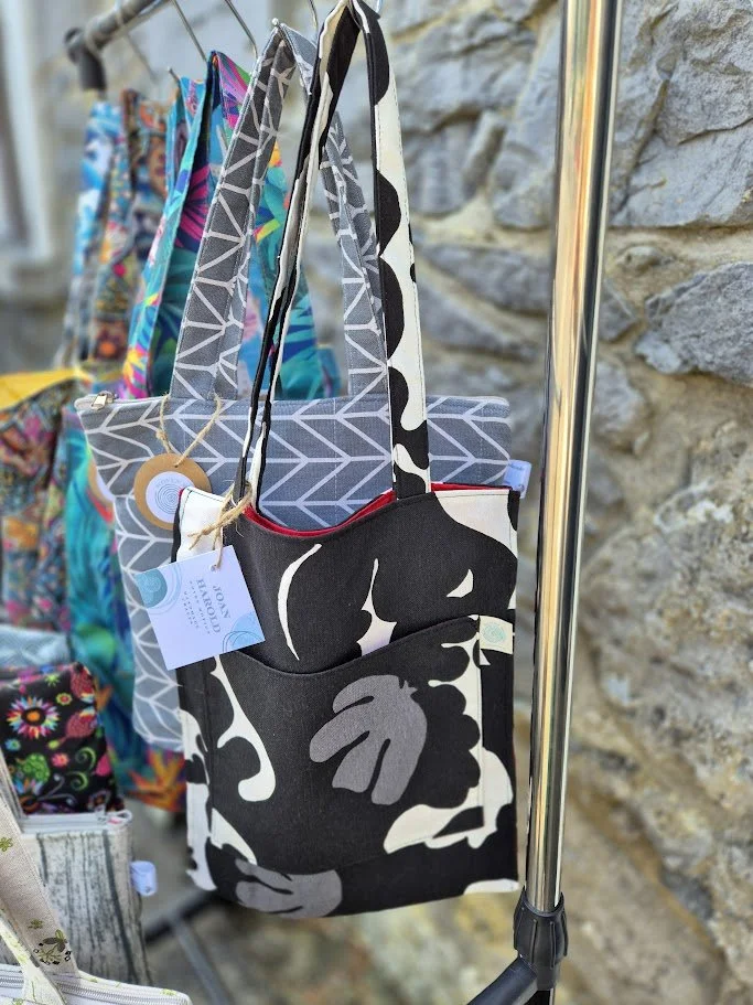 Colorful fabric tote bags hanging on a rack outside against a stone wall.