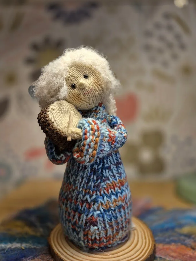 A knitted doll with white hair, holding a small woven accessory, standing on a wooden base.