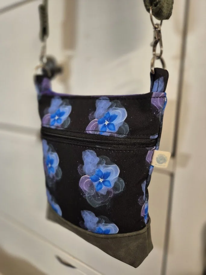 A black fabric bag with blue and purple flower patterns, hanging from a chain against a blurred background.
