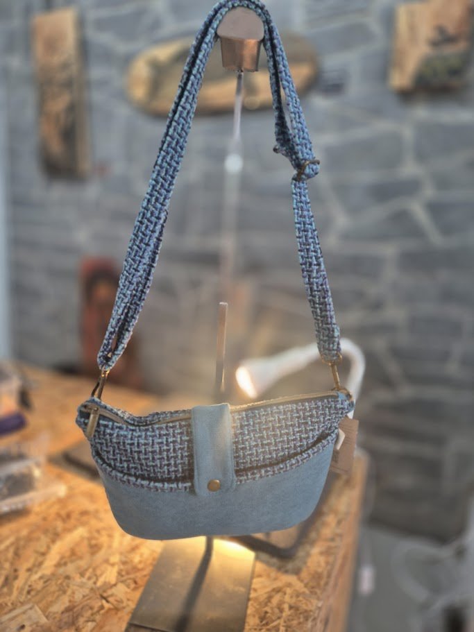 A small handbag with a mixed fabric pattern, hanging from a stand against a stone wall background.