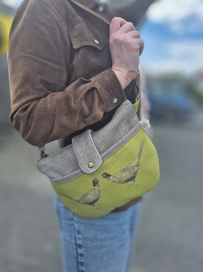 Person wearing a brown jacket and jeans, holding a gray and yellow bag with a bird illustration, outdoors with a blurred background.