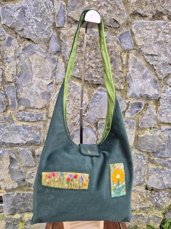 A green tote bag with two fabric patches, one with colorful flowers and the other with a yellow flower, hanging against a stone wall.