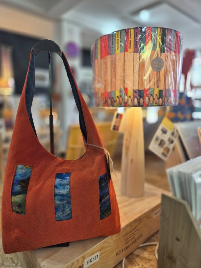 An orange handbag with four rectangular fabric patches that have a blue and green marbled pattern, displayed on a wooden shelf in a store. Behind it, a lamp with a colorful, patterned lampshade and a wooden base is visible.