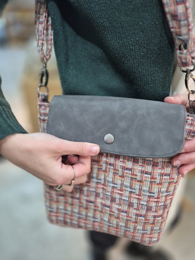 Person holding a gray clutch bag with a round metal clasp, wearing a multicolored plaid skirt, dark green sweater, and a ring on their finger.