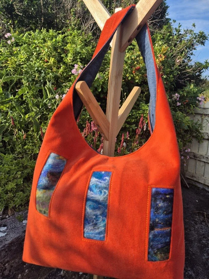 Bright orange fabric tote bag with three vertical fabric patches featuring abstract and nature-inspired designs, hanging on a wooden stand outdoors with green foliage and pink flowers in the background.
