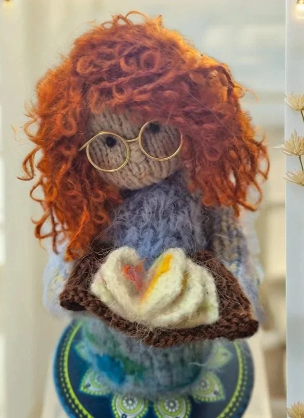 A handmade doll with curly red hair, round glasses, and gray outfit, holding a small wrapped food item.