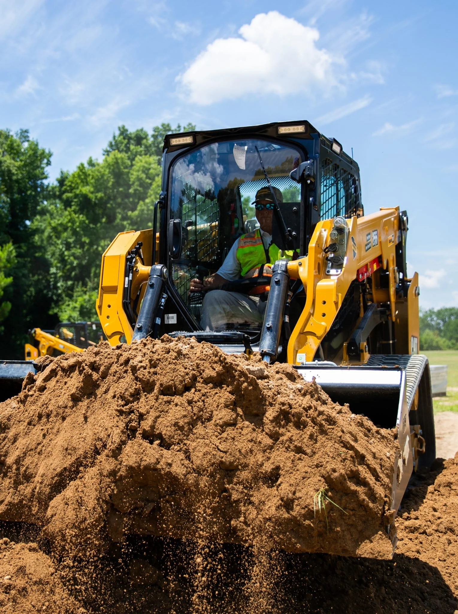 A construction worker operating a yellow bulldozer on a dirt field under a blue sky with clouds, surrounded by green trees.