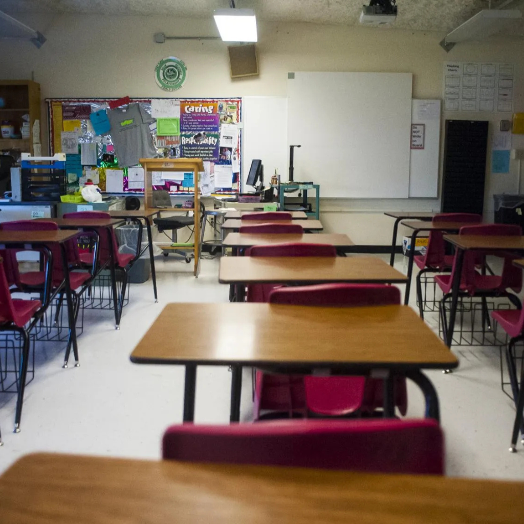 Empty classroom with desks and chairs, whiteboard at the front, bulletin boards with papers, and classroom supplies.