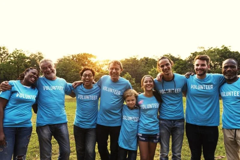 Group of diverse volunteers wearing matching blue shirts with 'VOLUNTEER' written on them, standing outdoors in a field at sunset, smiling and embracing each other.