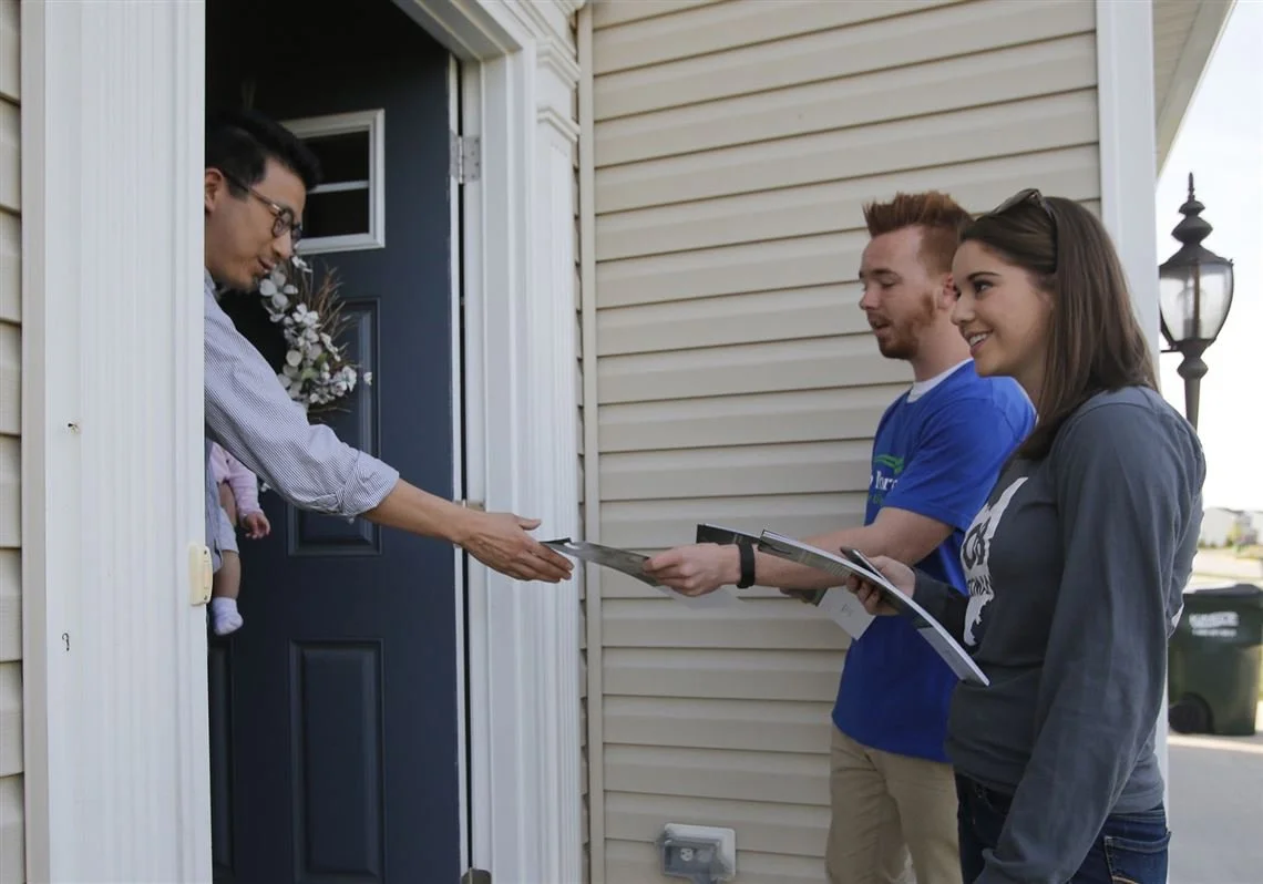A man at the door of a house handing an envelope to a couple who are standing outside, with the woman holding a clipboard.