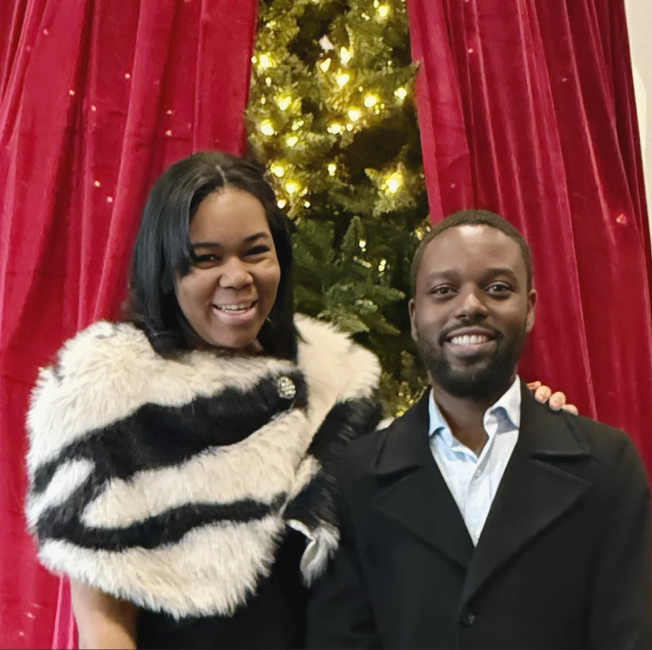 Jayla Thomas and her husband posing in front of a decorated Christmas tree with red curtains in the background.
