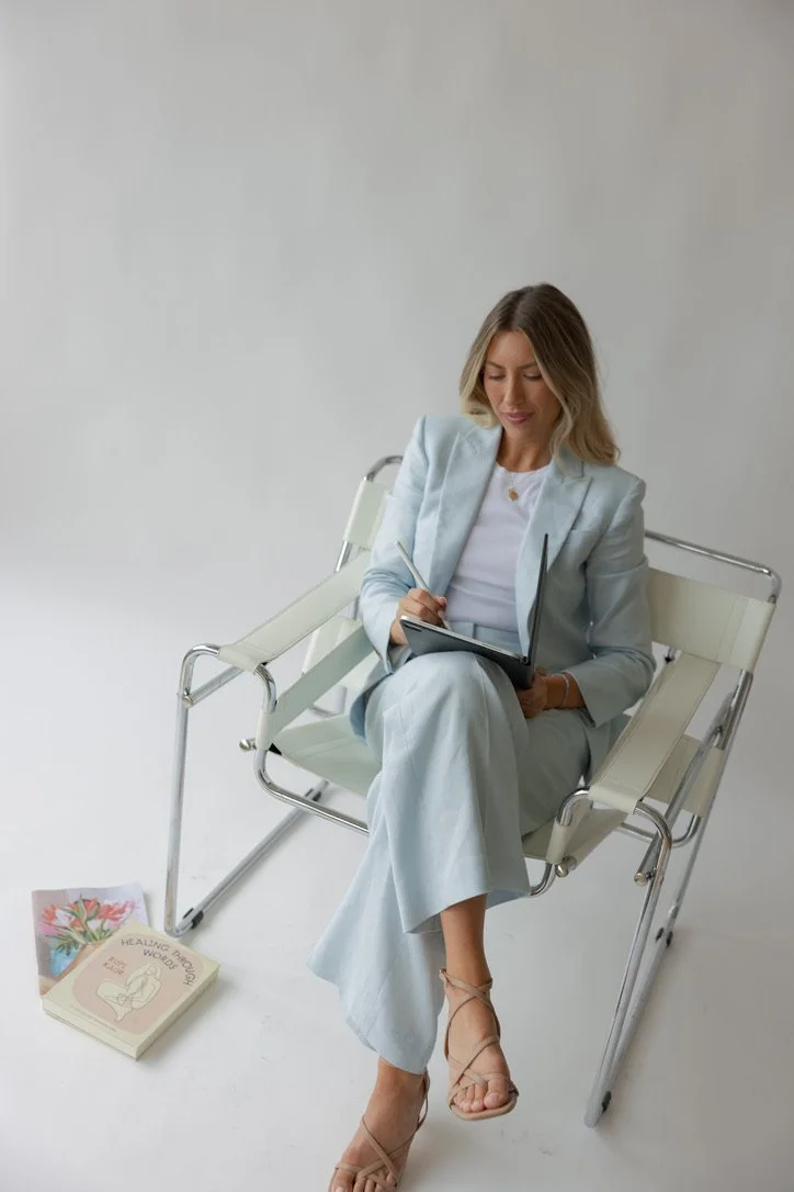 A woman in a light blue blazer and pants sitting on a modern white and chrome chair, writing in a notebook with a pen, in a minimalistic white studio space, with books on healing and flowers on the floor nearby.