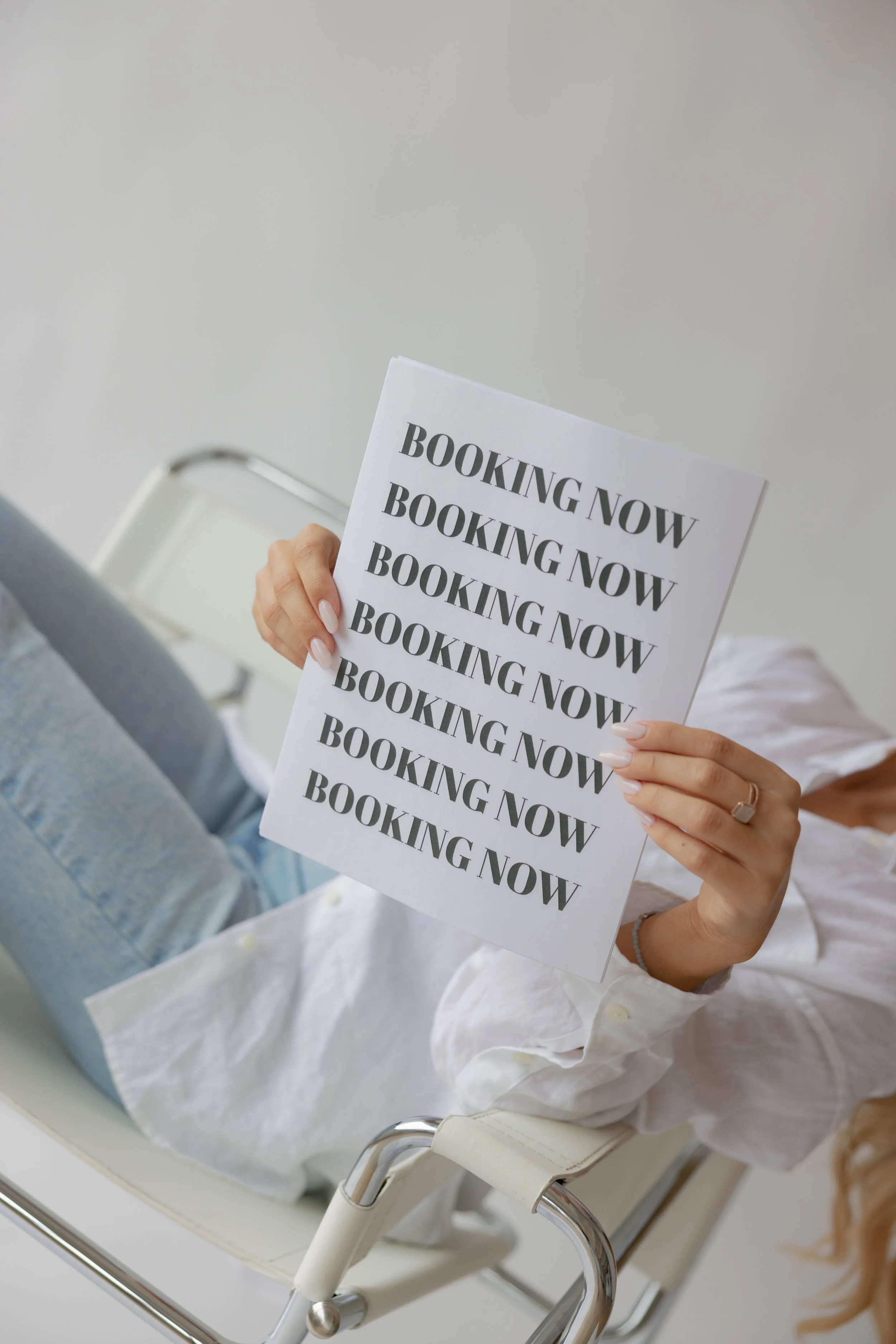 Person lying on a hospital bed holding a sign that repeatedly says 'Booking Now'