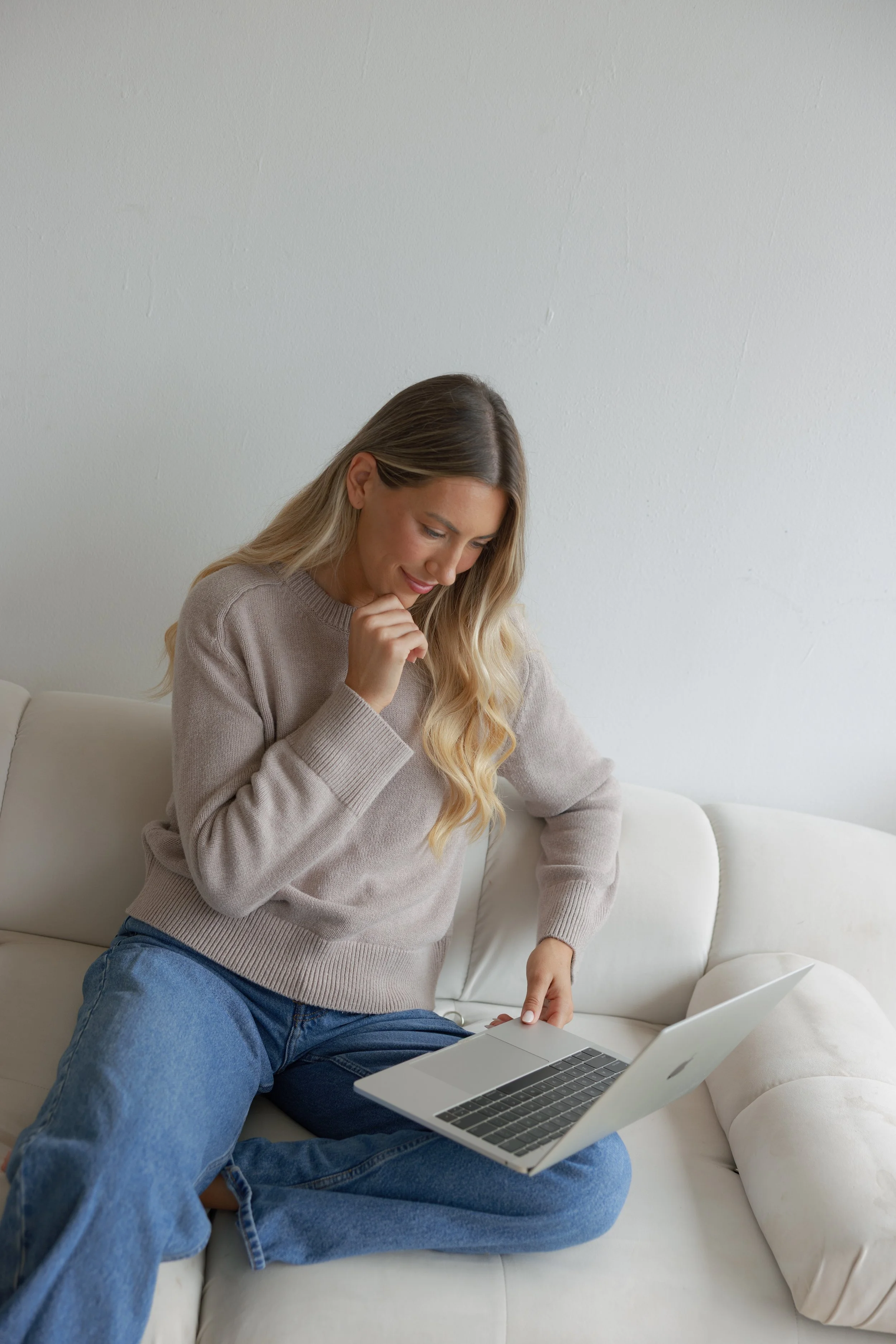 A woman sitting on a white sofa with a laptop, looking at the screen with a thoughtful expression.