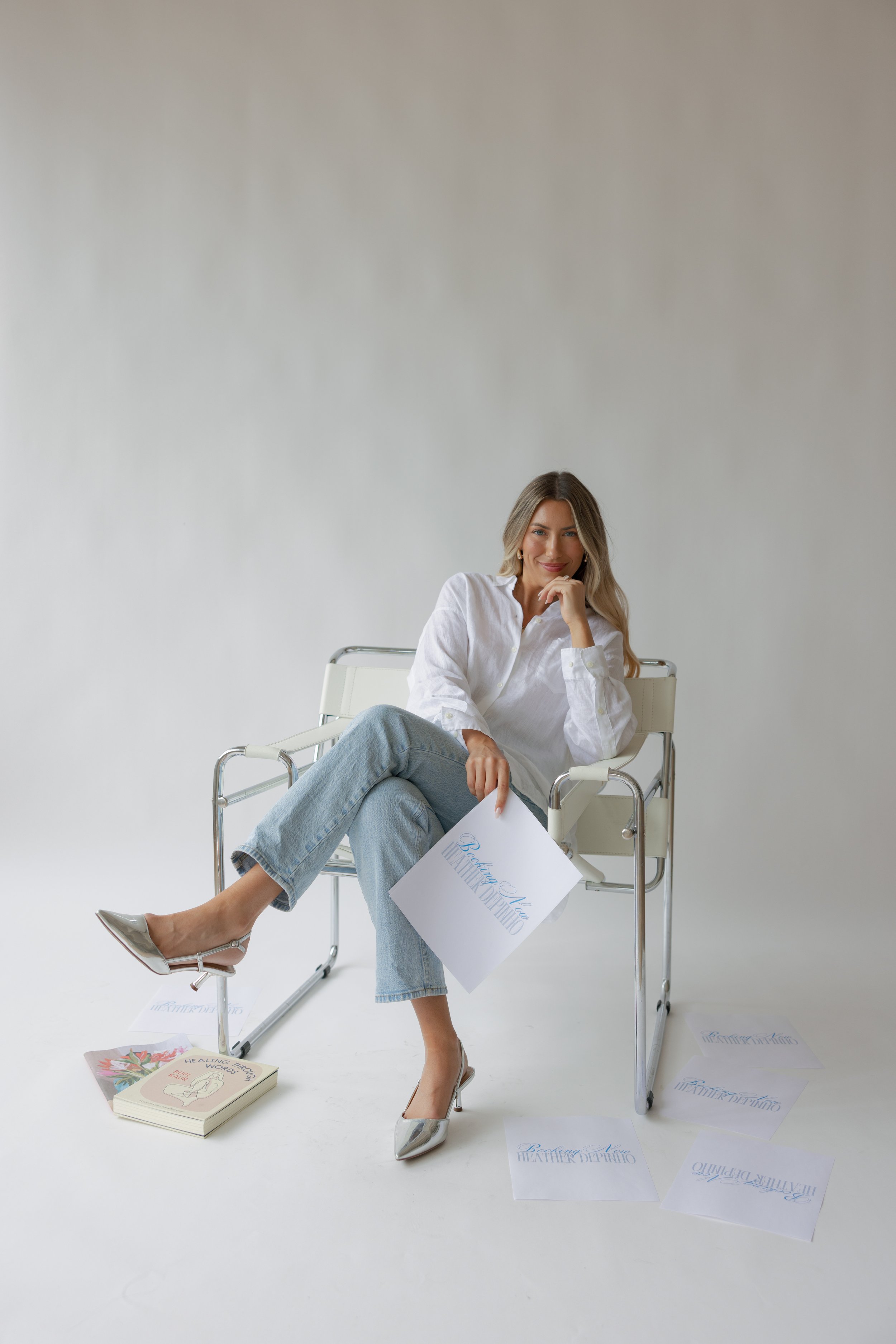 A woman sitting on a white chair, holding a paper with the words 'Beautiful You'. She has blonde hair, is wearing a white blouse, jeans, and silver heels, and is smiling at the camera. There are books and papers scattered on the floor around her, with a plain light background.