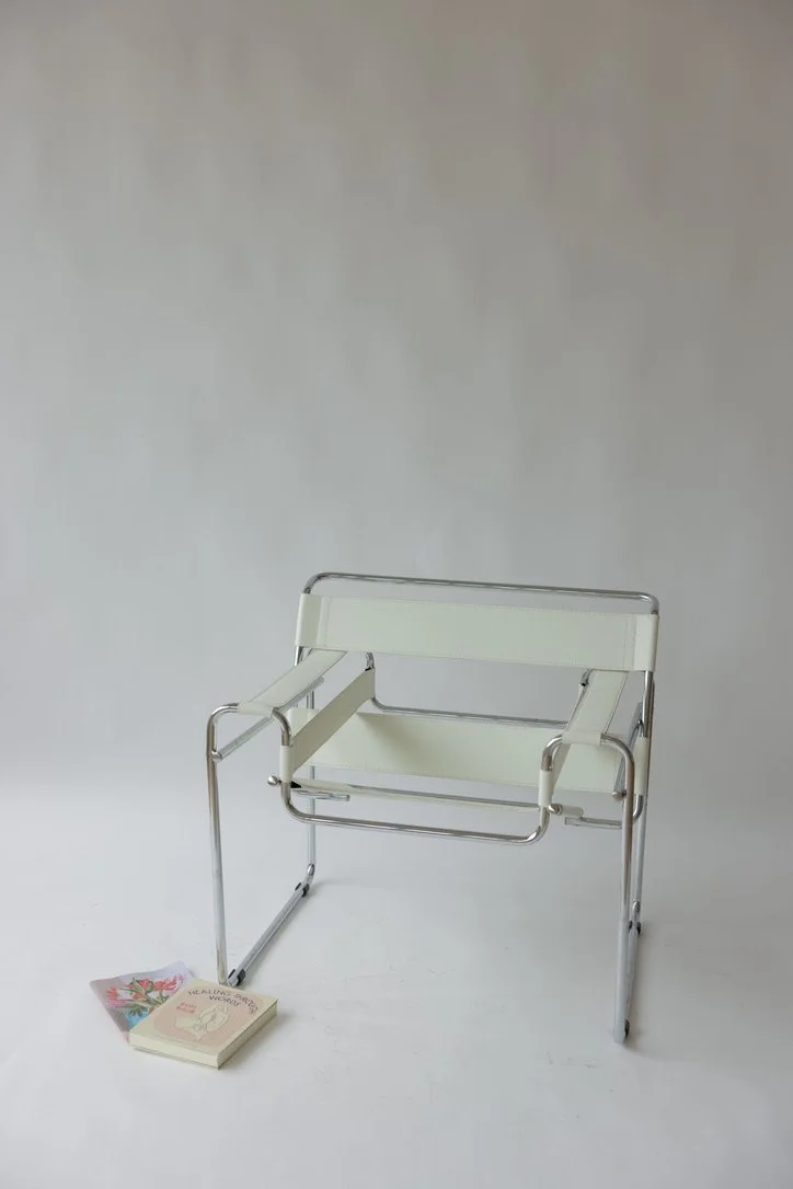 A white chair with a metal frame placed against a plain background, with a small book and a floral napkin on the floor next to it.