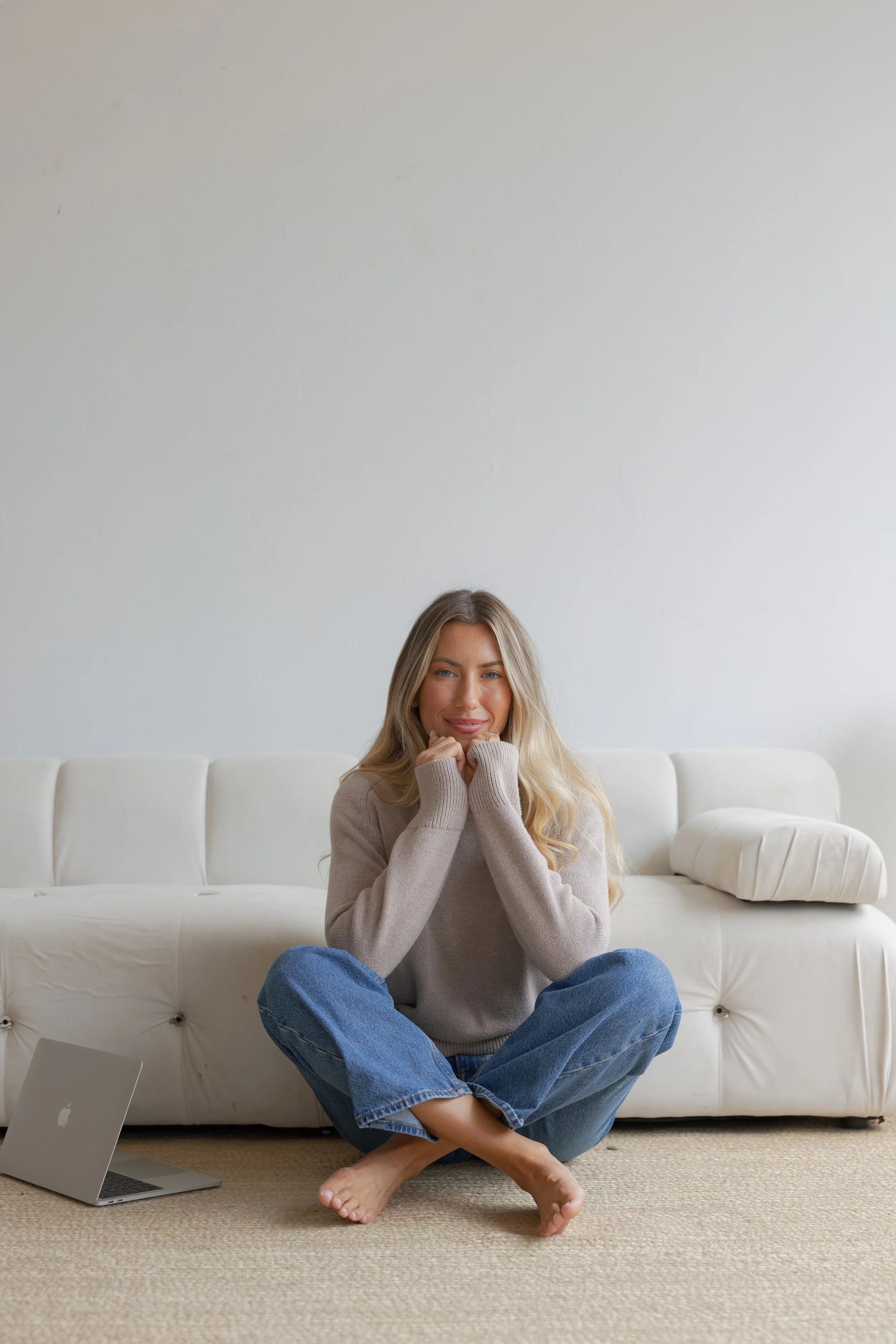 A young woman with long blonde hair sitting cross-legged on the floor in front of a white sofa, smiling, with a silver MacBook on the floor beside her in a bright, minimalistic room.