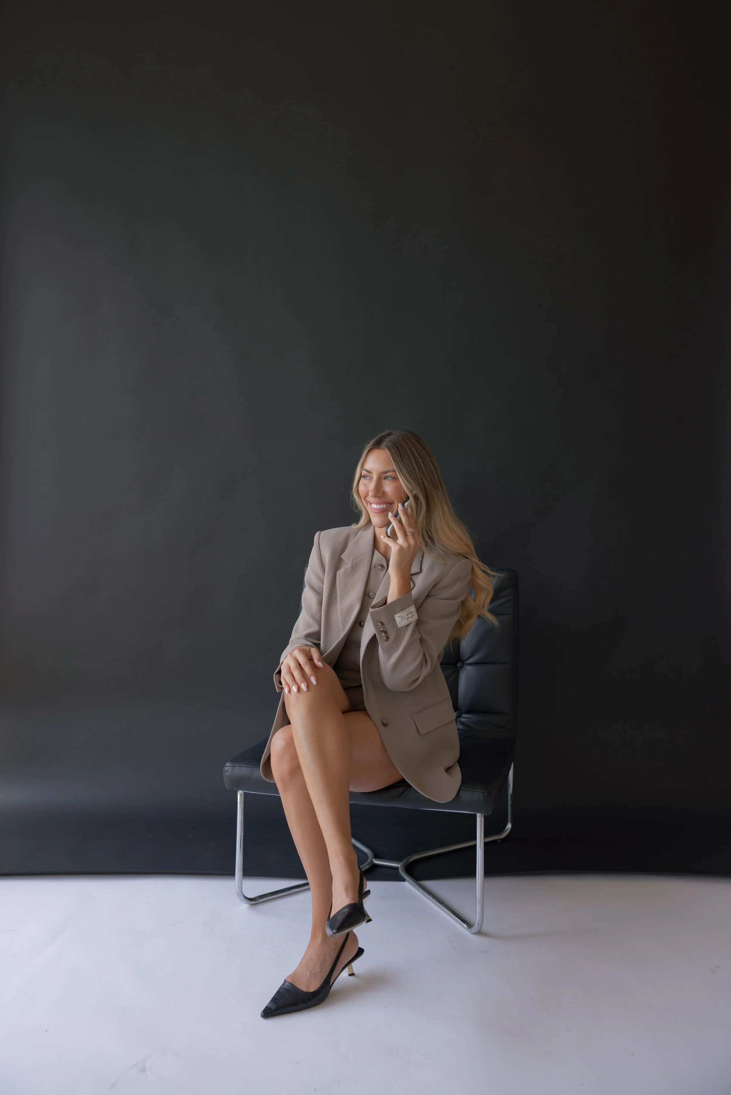 A woman in a beige blazer and high heels sitting on a black chair, smiling and talking on her phone, against a plain dark background.