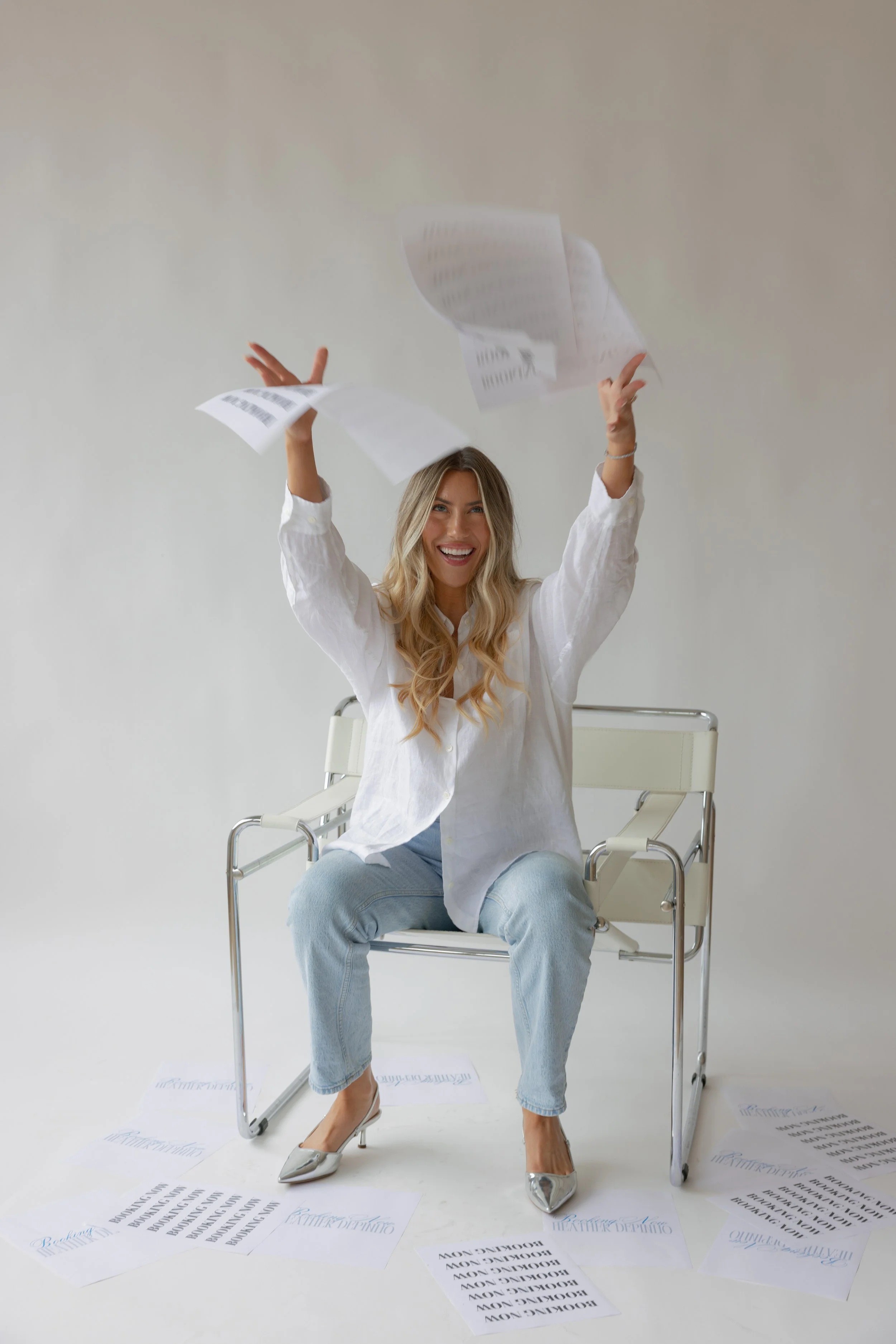 A woman sitting on a white chair, smiling, with papers falling around her and in her hands, wearing a white shirt, light blue jeans, and silver high heels.