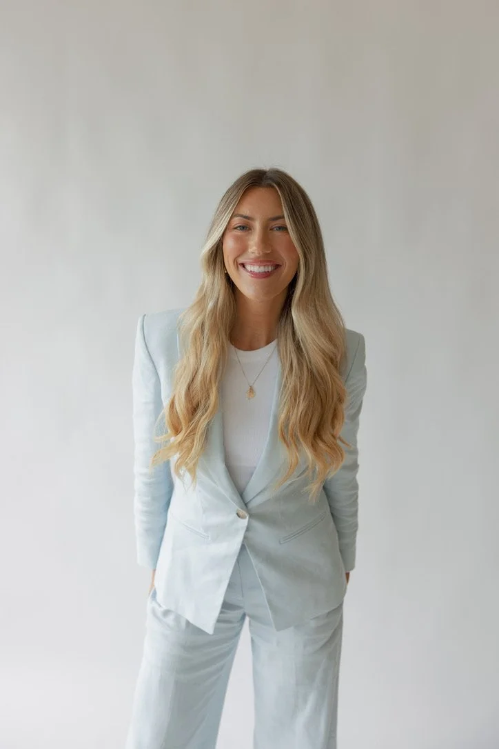 A woman in a light blue suit smiling, standing against a plain light background.