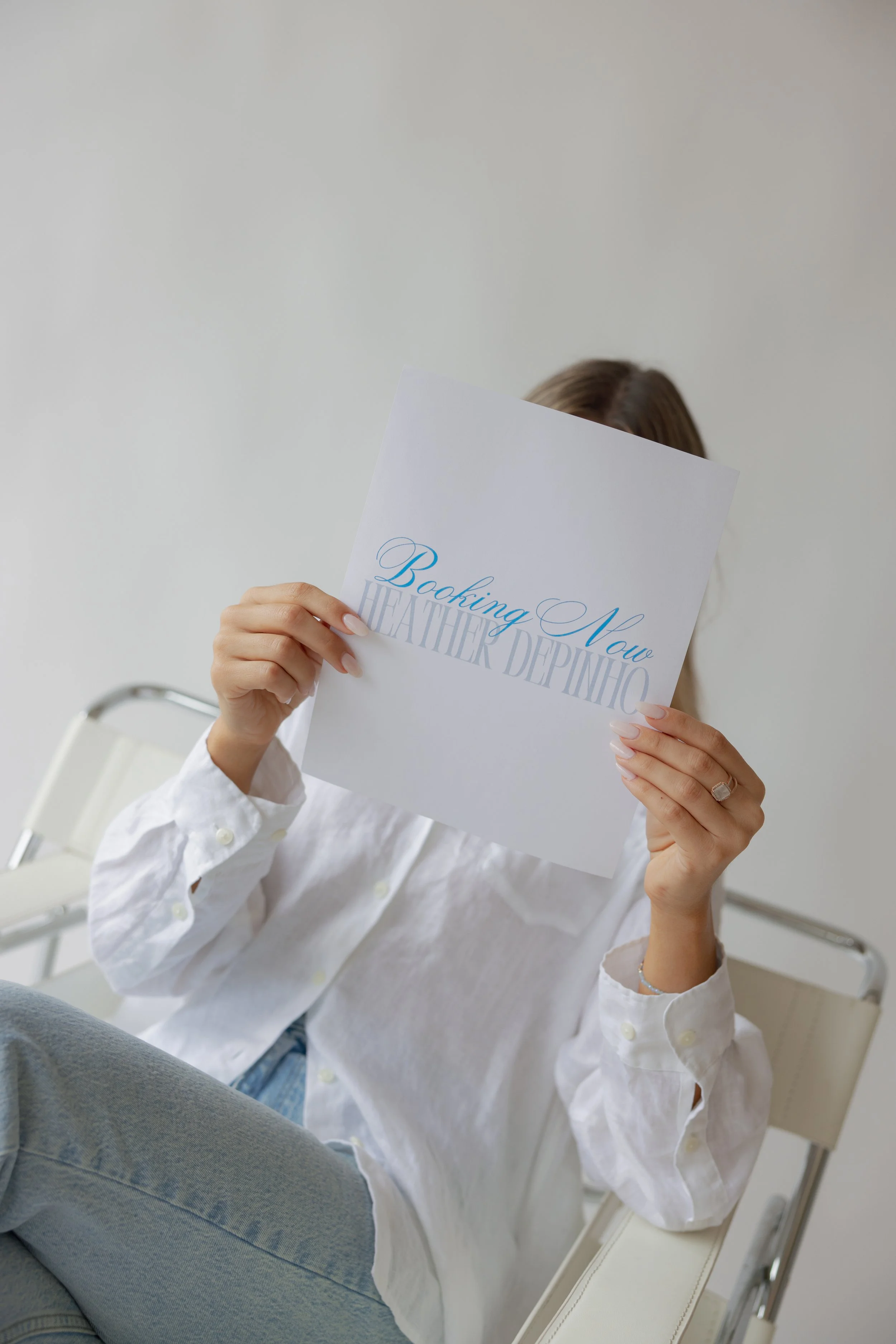Person holding a brochure that reads 'Booking Now WEATHER DEPINHO' while sitting on a chair, wearing a white shirt and light blue jeans.
