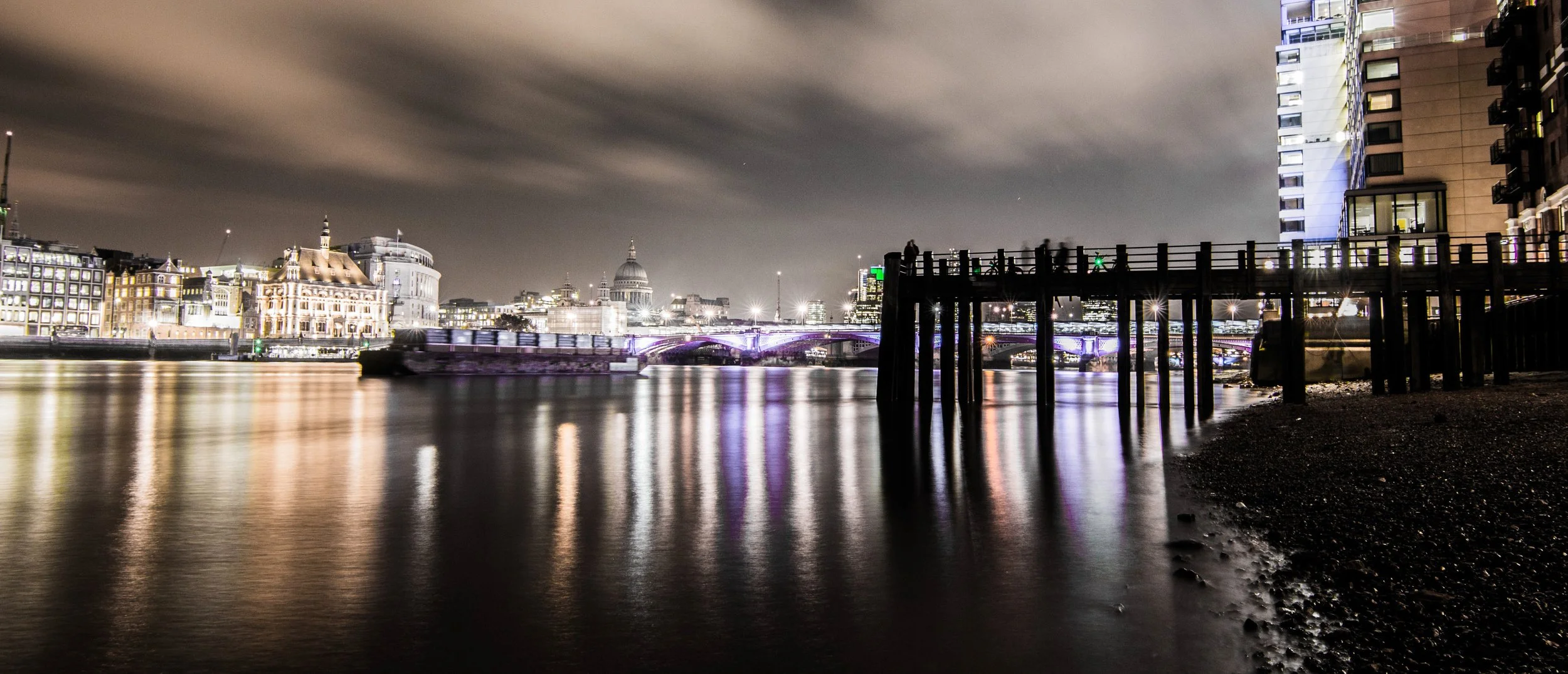 Nighttime cityscape of London featuring illuminated historic and modern buildings along the River Thames, with a bridge lit up in purple and a dock with wooden posts in the foreground.