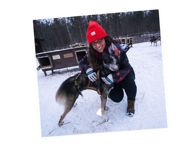 A woman smiling and hugging a dog outdoors in a snowy area, wearing a red beanie, glasses, and winter clothing.