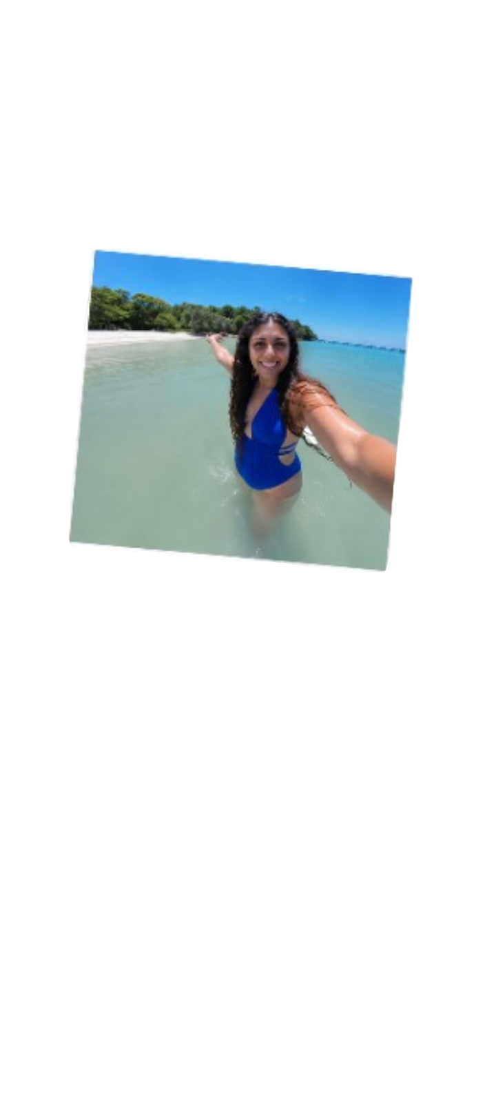 A woman in a blue swimsuit taking a selfie in the ocean, with a sandy beach and green trees in the background.