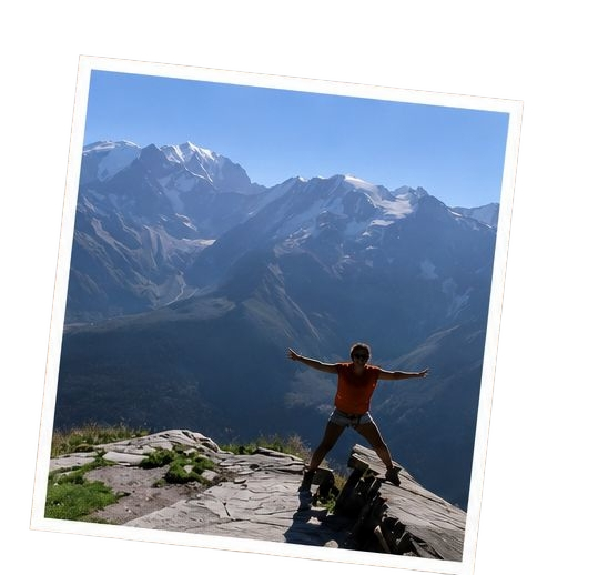 Person standing with arms outstretched on a mountain ledge with snowy mountain peaks in the background.