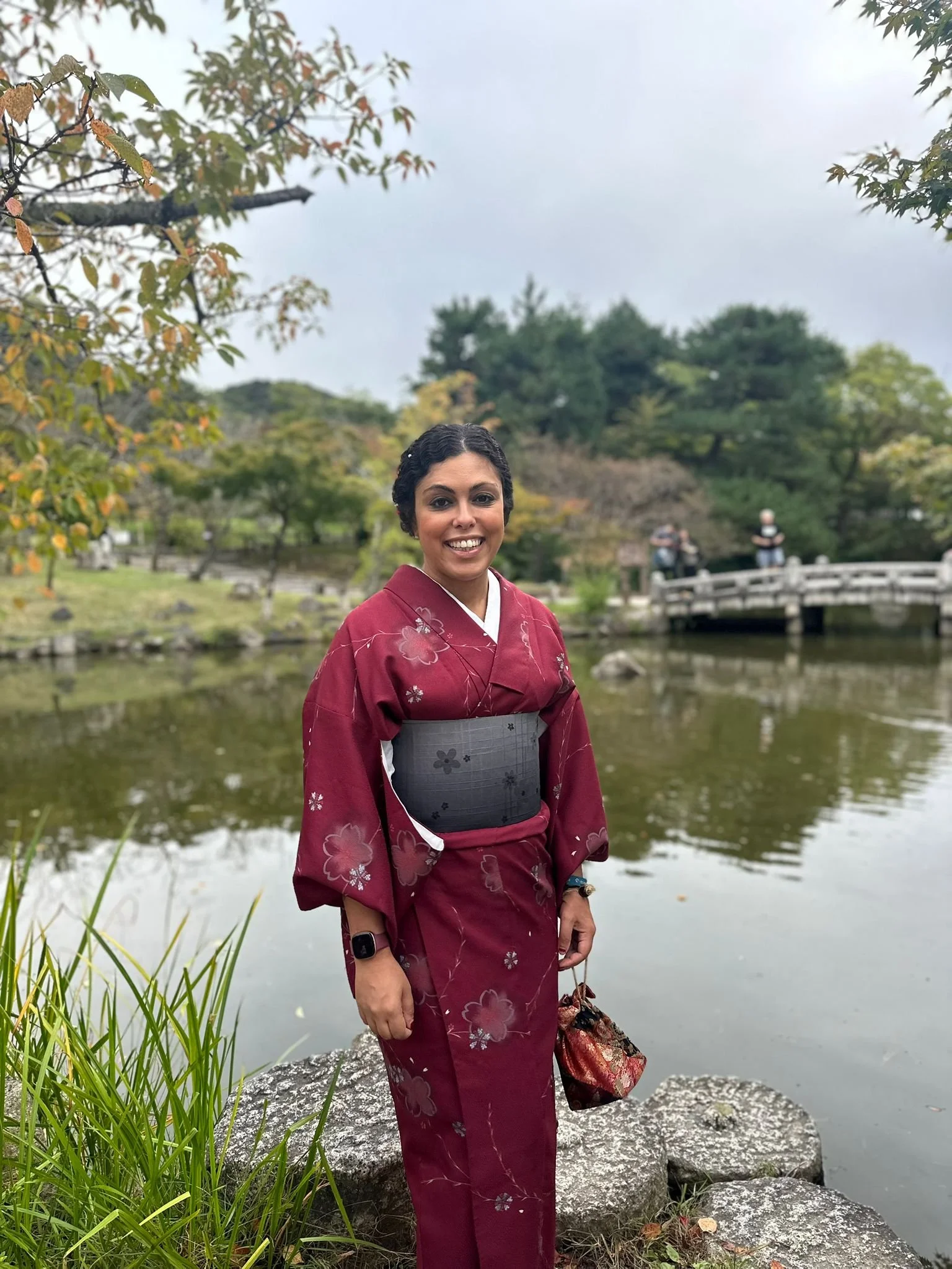 A woman in a maroon kimono with floral designs stands by a pond in a Japanese garden during fall, holding a small bag and smiling.