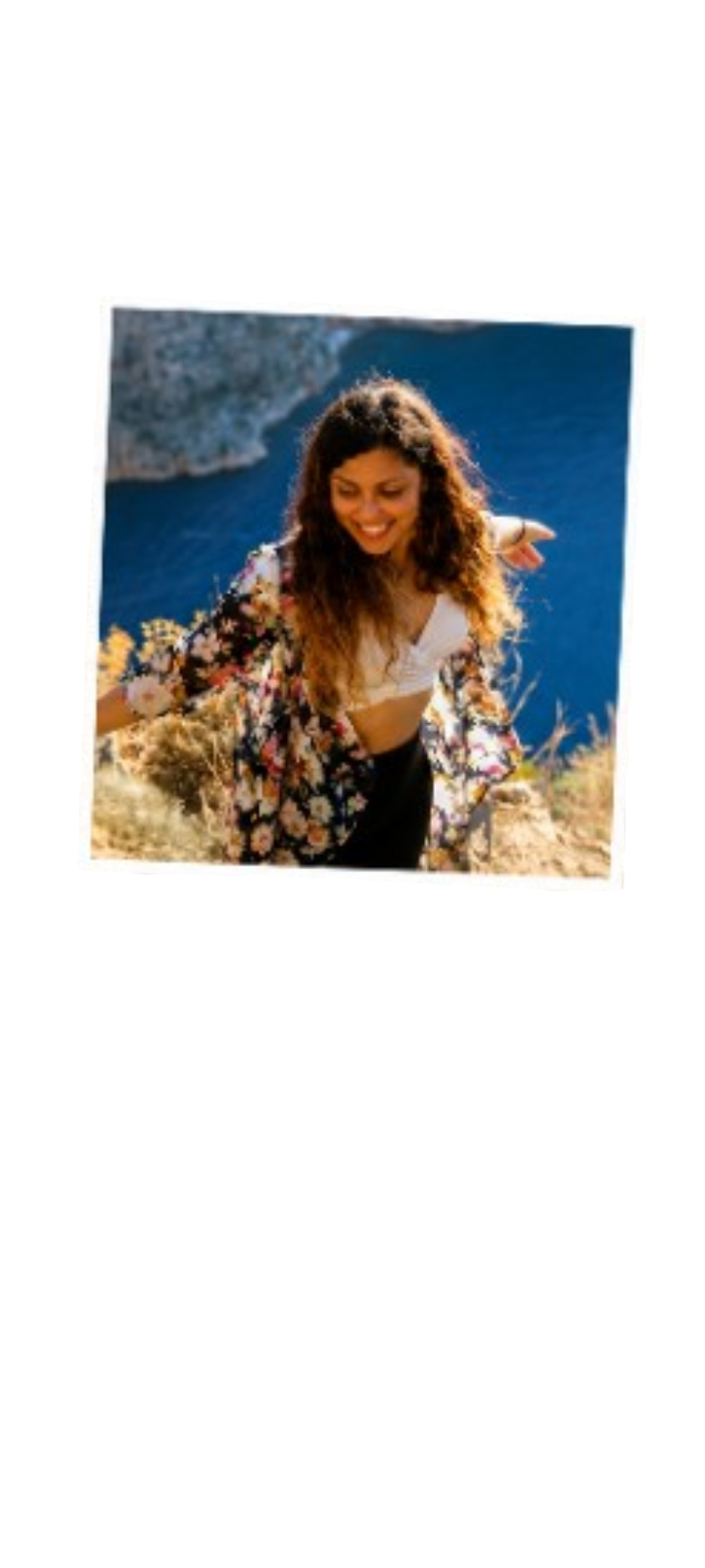 A woman with curly hair smiling and walking along a coastal trail with ocean waves behind her.