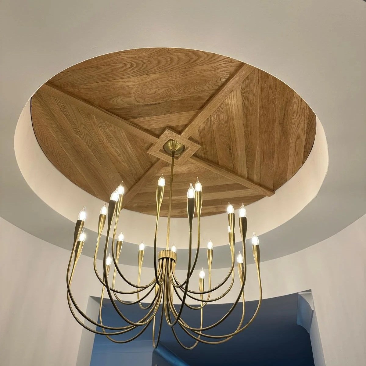 A modern chandelier with multiple gold-colored curved arms and candle-shaped light bulbs hanging from a ceiling with a circular wooden ceiling feature.