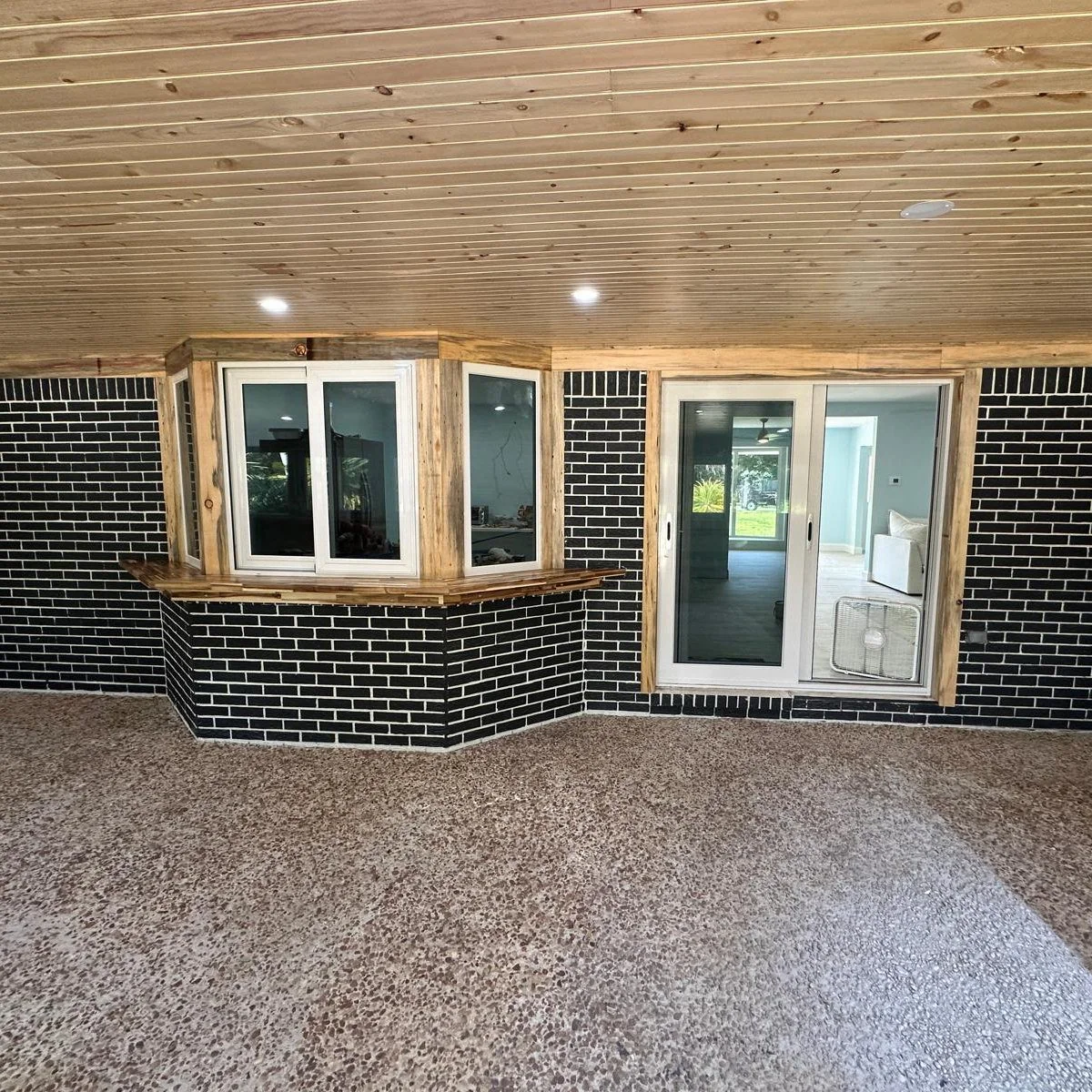 Interior view of a room with a textured brown floor, black brick wall accents, a wood-paneled ceiling, and large windows and sliding glass door.