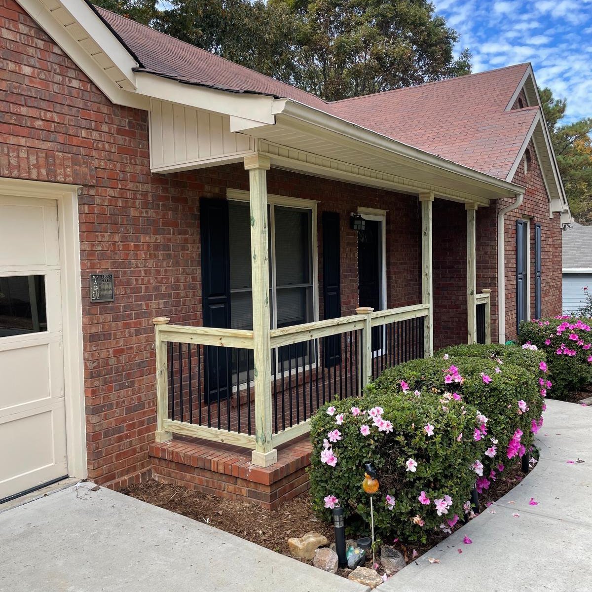 Front porch of a brick house with a small new wooden railing, black shutters on windows, and pink and white flowers in bushes along the sidewalk.