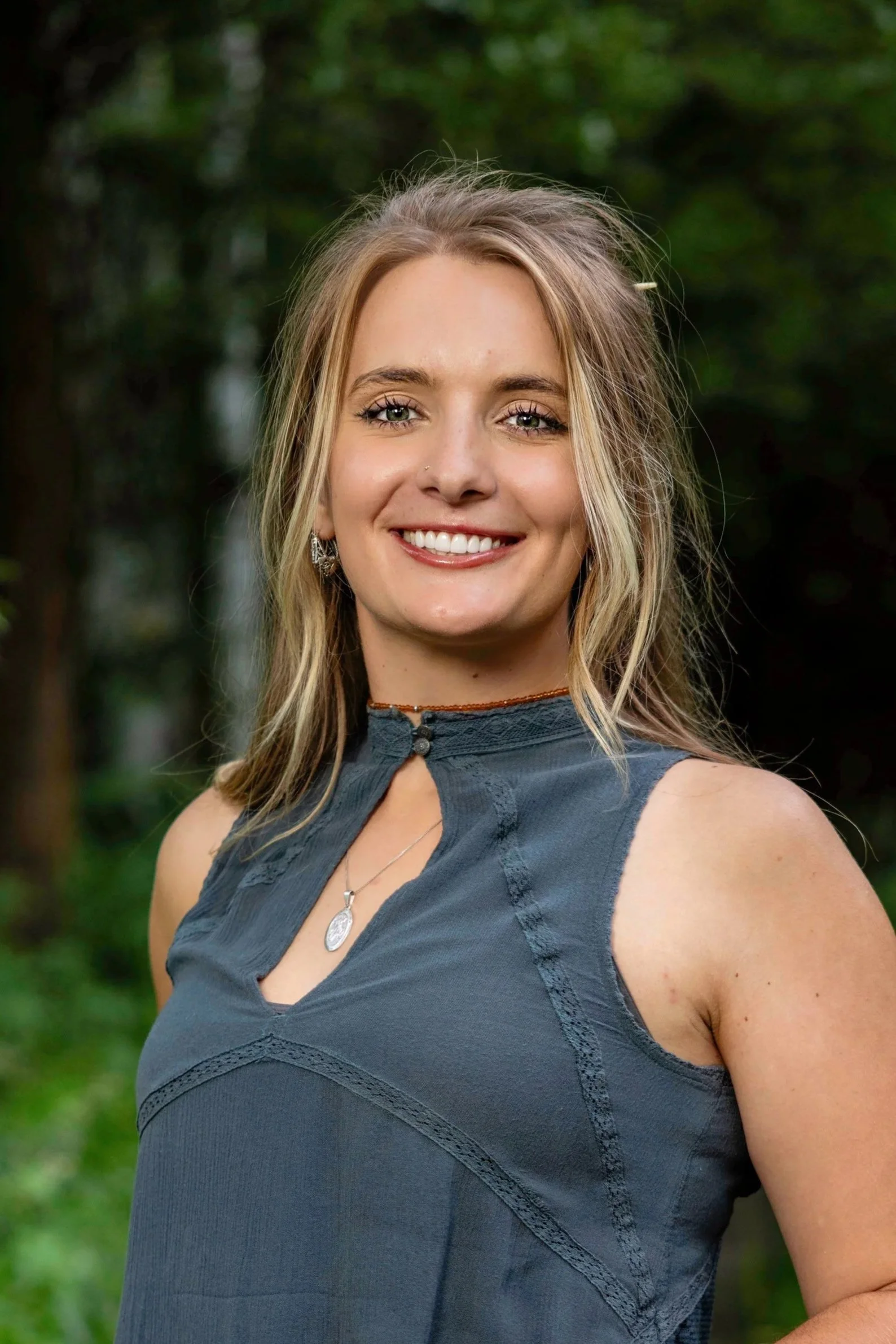 Portrait of a woman with blonde hair, smiling, wearing a sleeveless blue top, standing outdoors with green trees in the background.