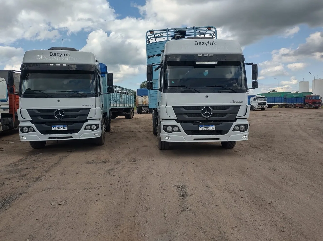 Dos camiones Mercedes-Benz de color gris estacionados en un terreno de tierra, con otros camiones en el fondo y un cielo nublado.