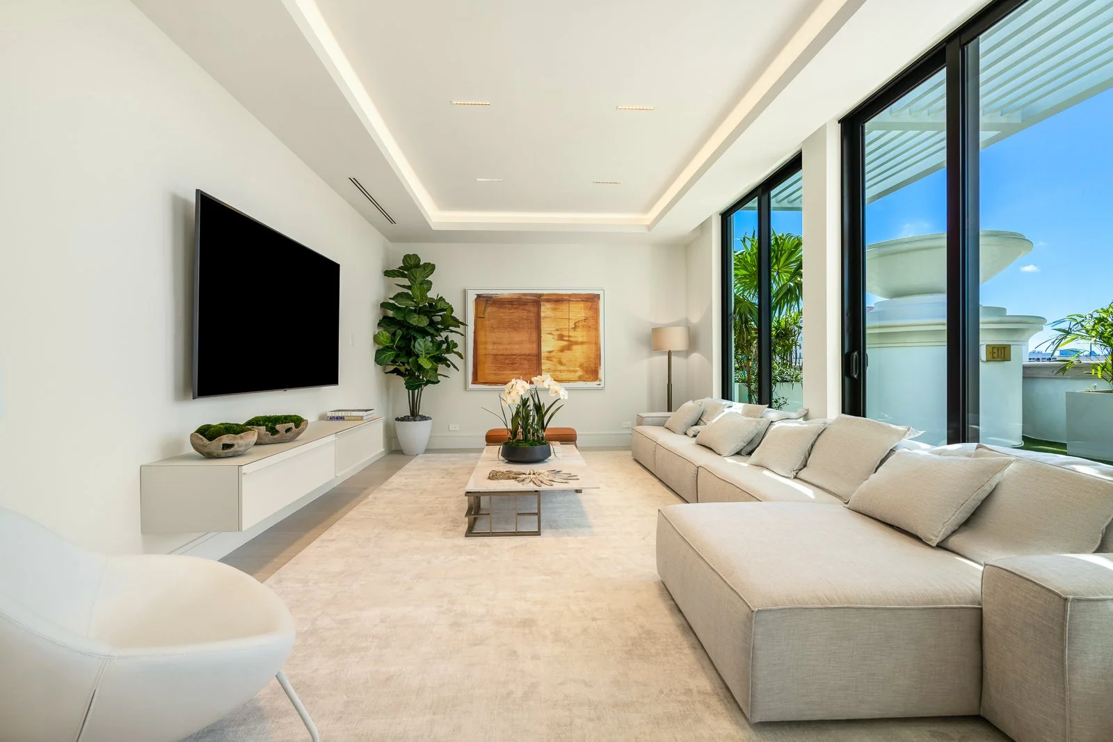 Bright living room with large beige sectional sofa, coffee table with plants, flat-screen TV on white wall, sliding glass doors with outdoor view, and tropical plants outside.