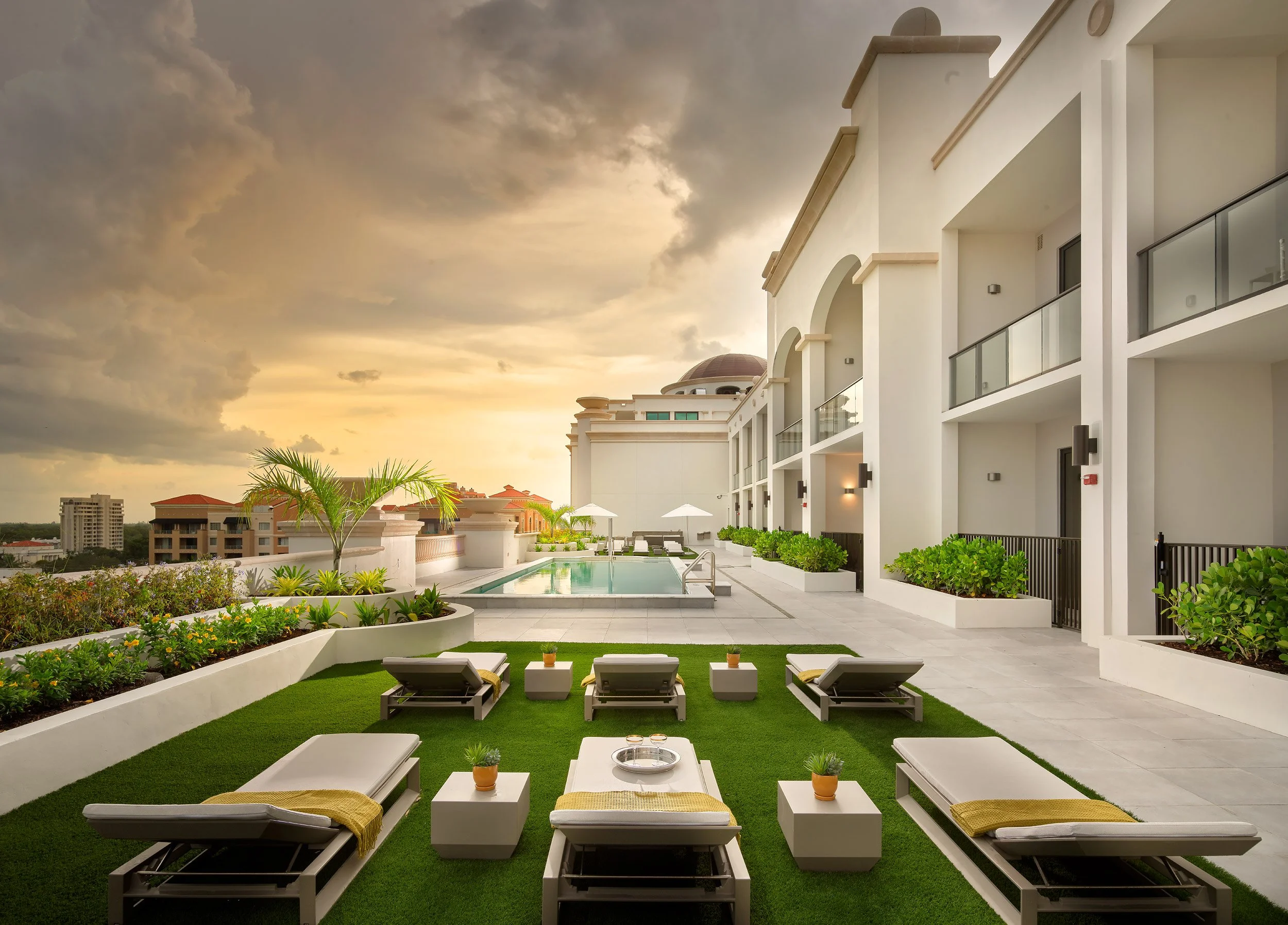 Luxury rooftop terrace with swimming pool, lounge chairs, potted plants, and green landscaping, overlooking city buildings at sunset.