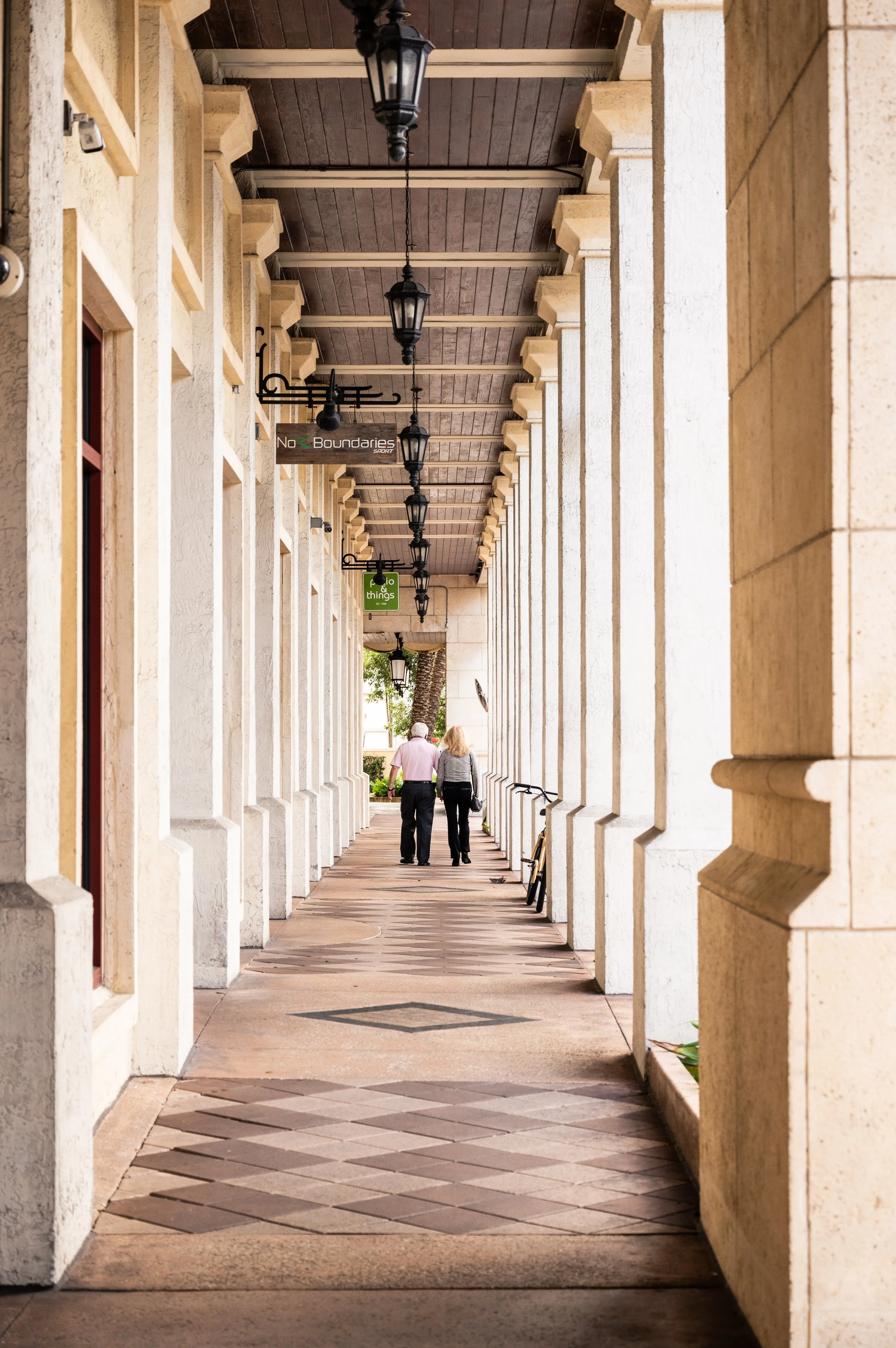 A man and a woman walking in an outdoor arcade or sidewalk with tall, beige columns on the right and left, hanging black lanterns overhead, and a sign that reads 'No Boundaries'.