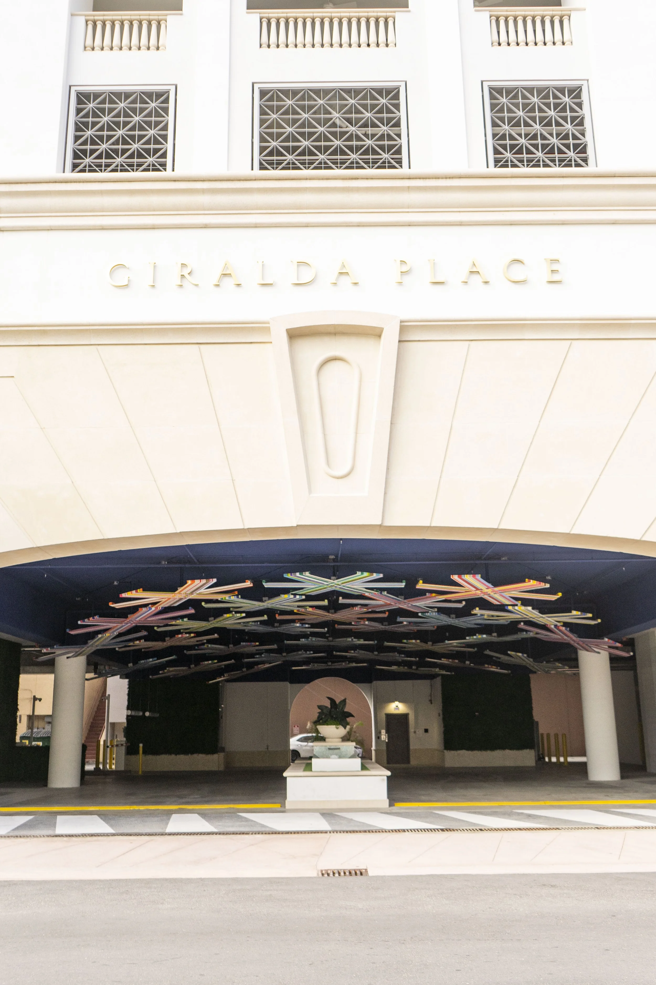 The image shows a building with the name 'GIRALDA PLACE' on the front. Below the name, there is an archway leading to an area with a decorative ceiling and a small white planter with green plants in the center.