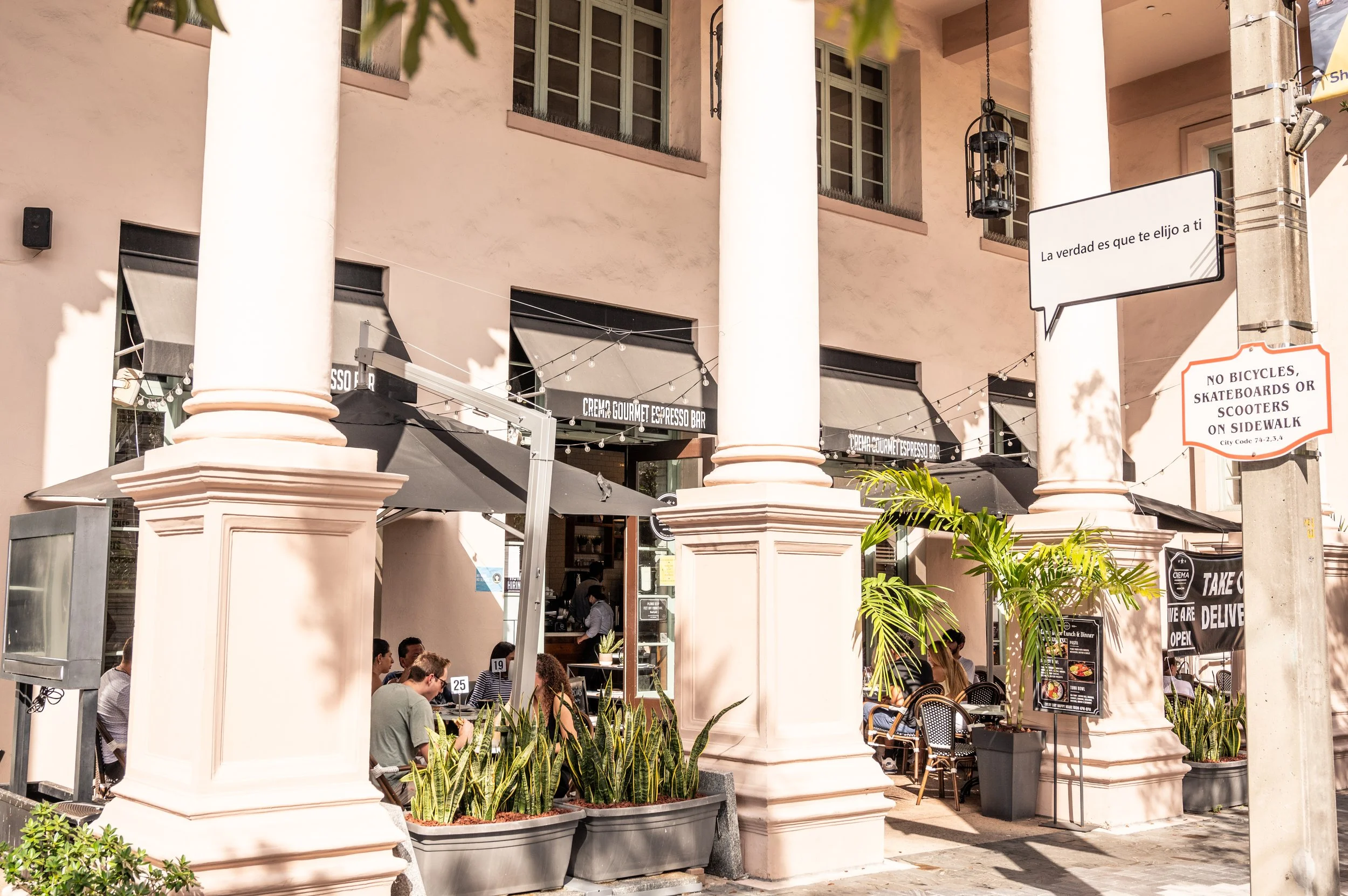 Outdoor seating area of a cafe or restaurant with patrons sitting at tables under umbrellas, pink building with large white columns, potted plants, and signs that include one in Spanish and one restricting bicycles and scooters on the sidewalk.