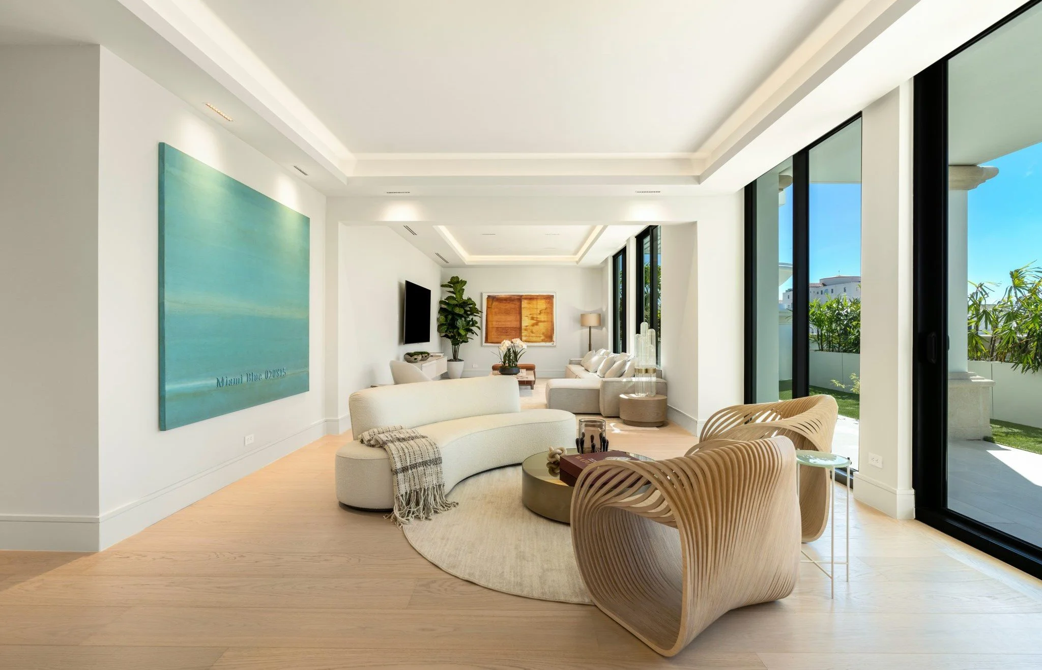 Modern living room with white walls, large glass sliding doors, contemporary furniture including a curved sofa, wooden chairs, and a round coffee table, with artwork and plants, and an outdoor patio visible.