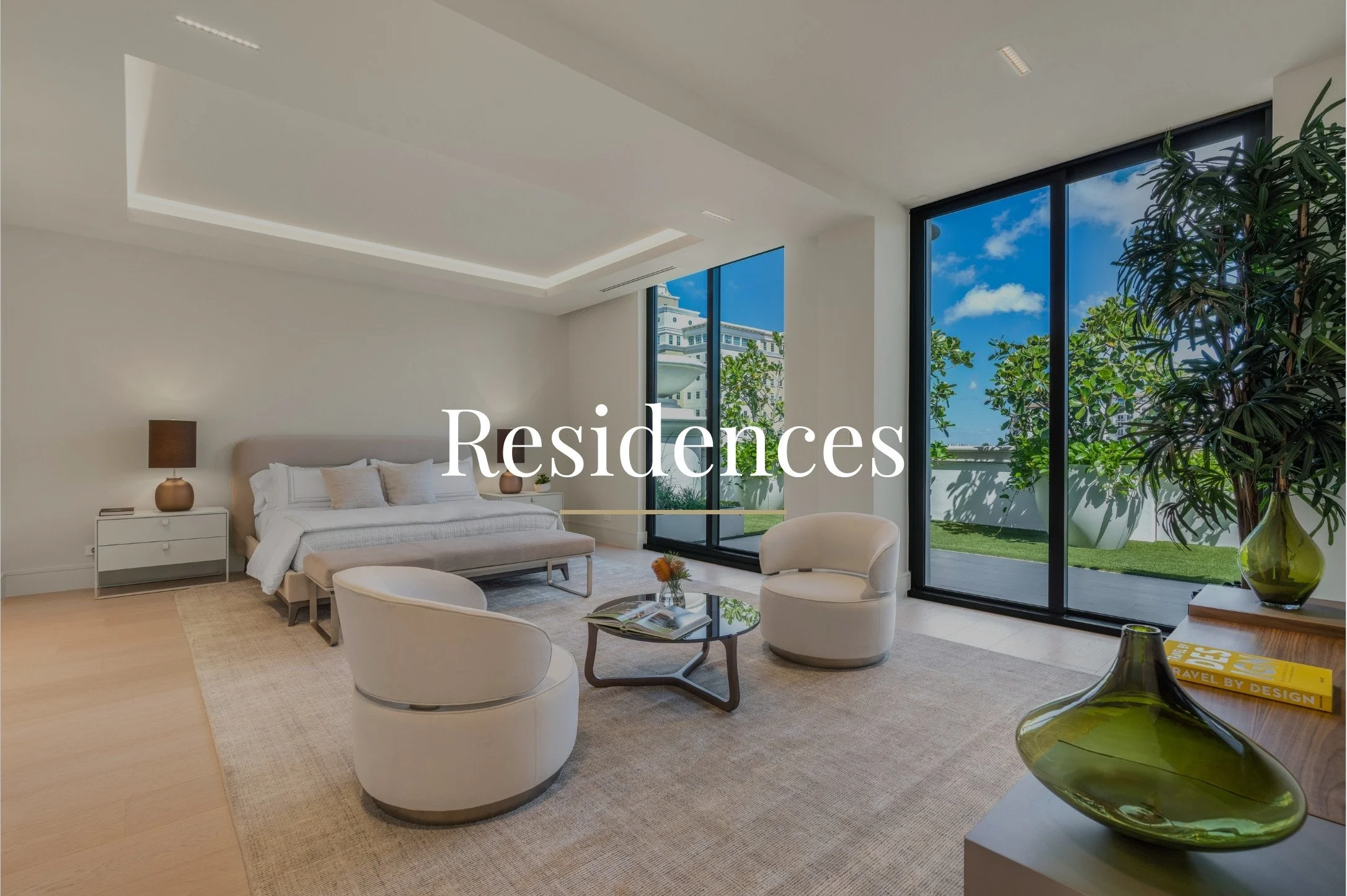 Modern living room with large floor-to-ceiling windows overlooking a green outdoor space, containing white furniture, a bed, chairs, a small table, and decorative items.