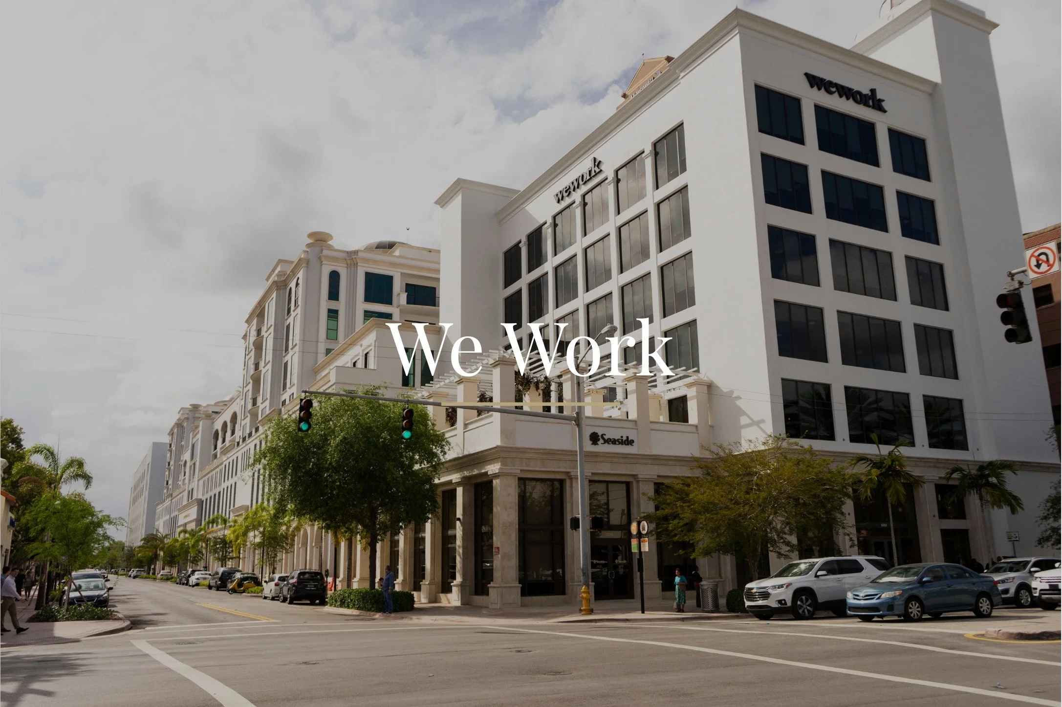 A city street with modern white buildings, parked cars, pedestrians, and green trees, under a cloudy sky. The words "We Work" are overlaid on the image.