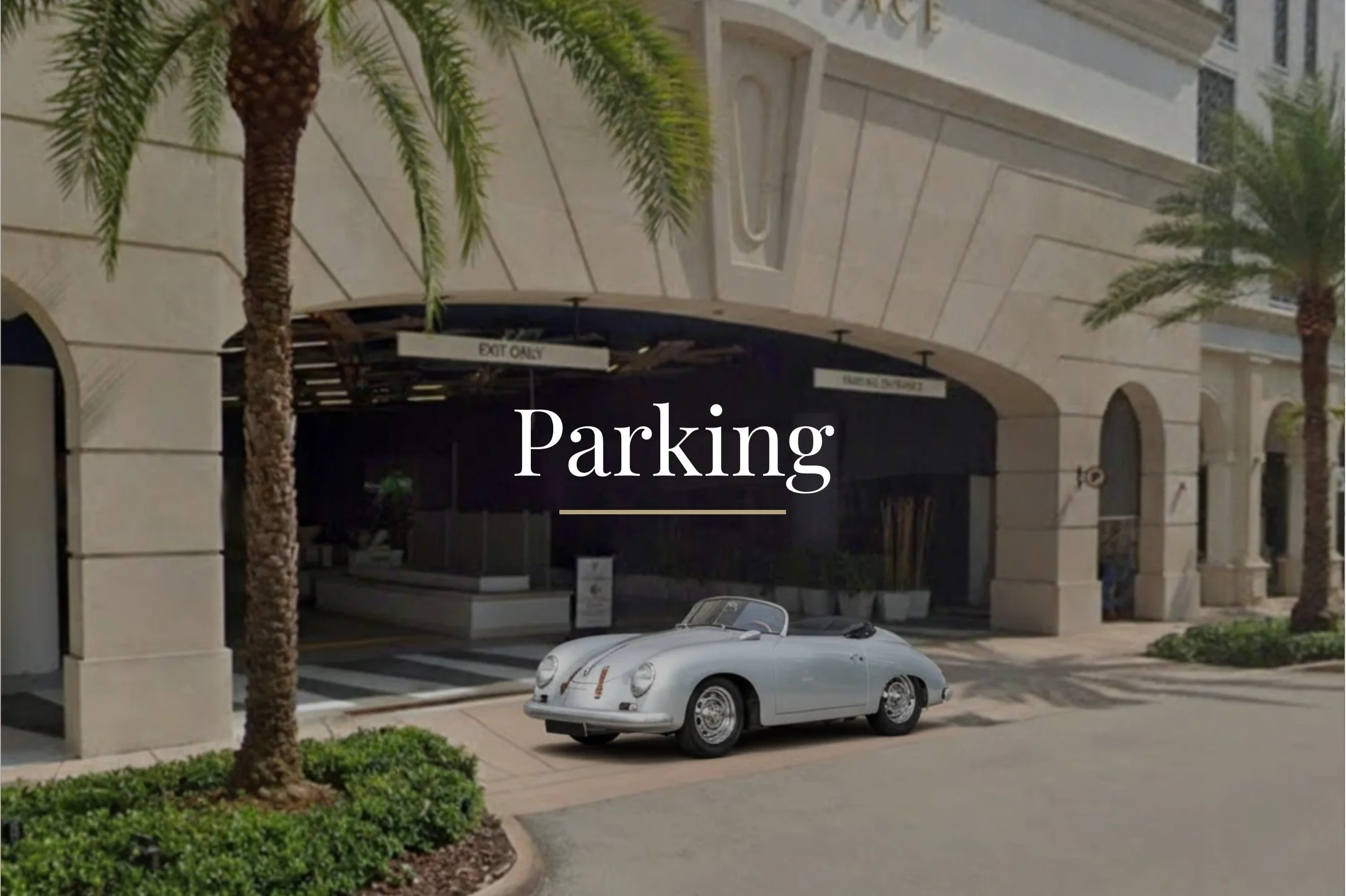 A vintage silver convertible Porsche parked in front of a building with palm trees, sidewalk, and an entranceway labeled 'EXIT ONLY' in a downtown or urban area.