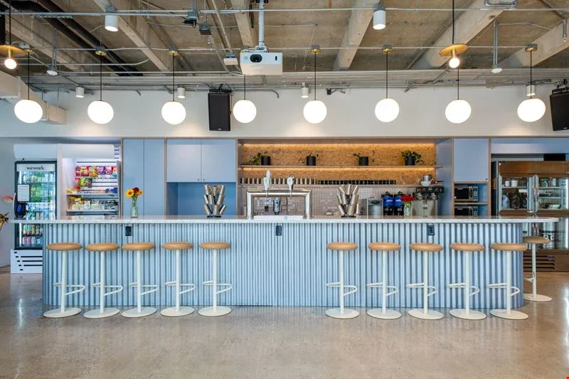 Modern bedroom with white walls, large sliding glass doors opening to a green outdoor space, beige bed with white bedding, two nightstands with brown lamps, two white round chairs, a glass coffee table, large green potted plant, and decorative items including books and vases.