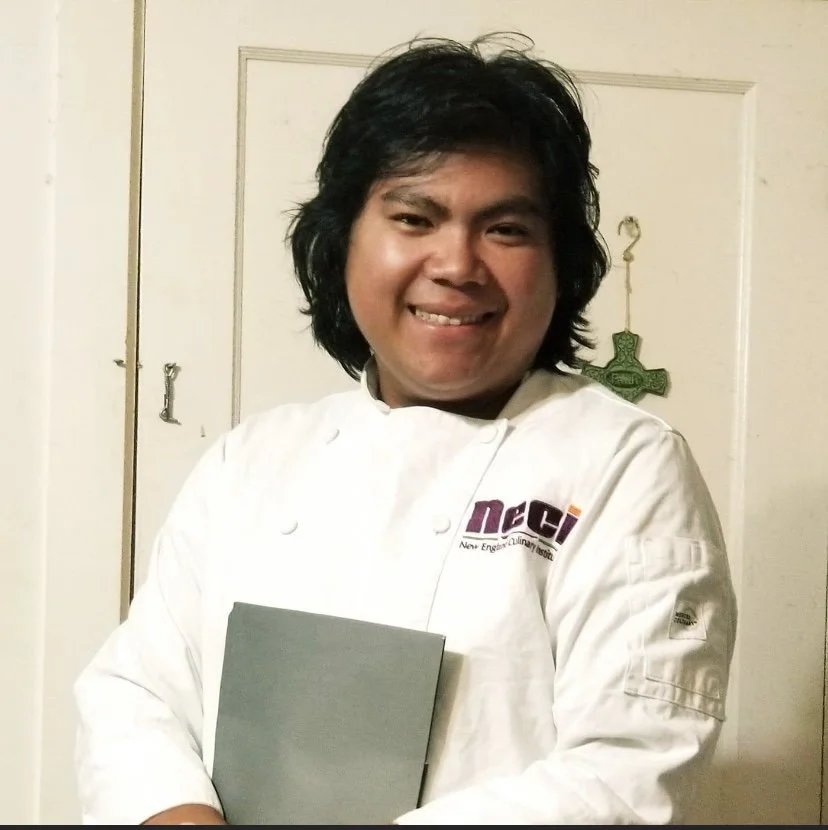 A smiling person with dark, shoulder-length hair wearing a white chef's jacket, holding a gray sheet of paper or notebook, standing in front of a white door with a decorative green cross hanging on it.