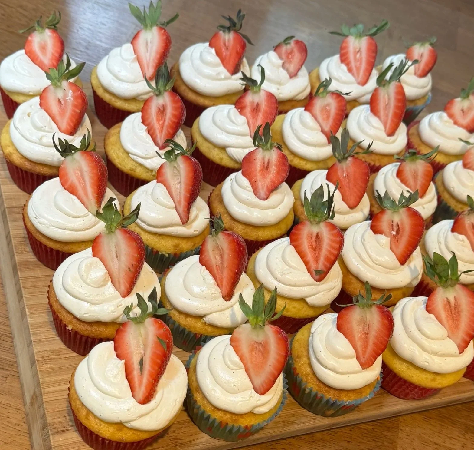 Tray of strawberry-topped cupcakes with white frosting arranged in colorful paper liners on a wooden surface.